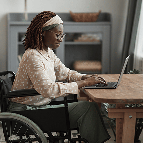 Woman sitting in wheelchair while leaning over desk to type on laptop